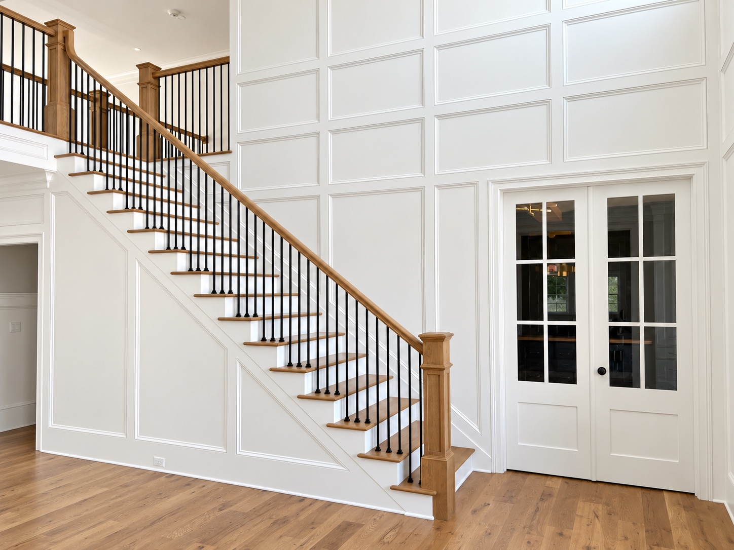 Finished staircase with paneled wood newels, black iron balusters, and white wall paneling surrounding glazed double doors